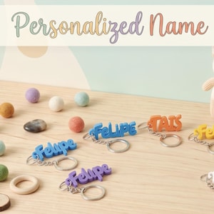 Könnte beinhalten: Eine Sammlung personalisierter Namensschlüsselanhänger in verschiedenen Farben wie Blau, Orange, Gelb, Weiß und Grün mit Metallringen. Der Text Personalized Name Keychains! steht über einem kleinen Plüschfuchs und drei gestapelten Würfeln.