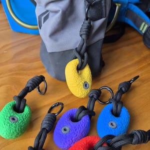 Peut inclure: Une collection de porte-clés en forme de prises d'escalade jaunes, vertes, violettes, bleues et rouges, avec une surface texturée, un trou central muni d'un œillet gris et une boucle en paracorde noire fixée à un petit mousqueton en métal noir.