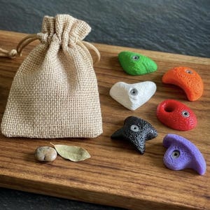 Puede incluir: Una bolsa de arpillera beige con cordón junto a seis pequeñas presas de escalada de colores verde, naranja, blanco, rojo, negro y morado, cada una con un orificio central de metal, además de un guijarro liso y una pequeña hoja seca.