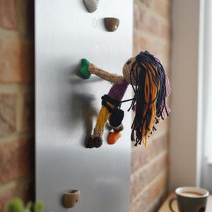 Può includere: Una bambola all'uncinetto con lunghi capelli di lana nera e arancione, che indossa un top viola e pantaloni gialli, è fissata a un pannello metallico verticale argentato. La bambola è in posa da arrampicata, aggrappata a piccole prese colorate.