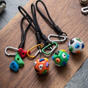 Peut inclure: Trois porte-clés avec des cordons noirs et des mousquetons en métal argenté, doré et vert. Deux porte-clés présentent des sphères géométriques multicolores avec des prises d'escalade, et un possède deux prises individuelles bleue et verte.