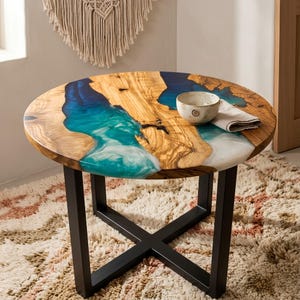 Pode incluir: Uma mesa redonda com superfície de madeira natural e uma incrustação de resina azul e turquesa em forma de rio. A mesa é sustentada por uma base de metal preto em forma de X. Uma pequena tigela de cerâmica com espiral está sobre o tampo.