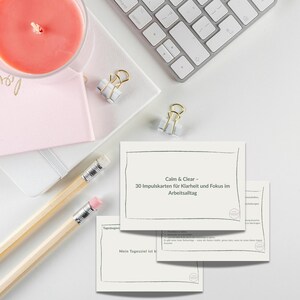 Peut inclure: Une scène de bureau avec une bougie rose allumée, des cahiers, des crayons, des trombones dorés et un clavier. Plusieurs cartes avec le texte "Calm & Clear" et du texte allemand sont visibles, suggérant la clarté et la concentration.