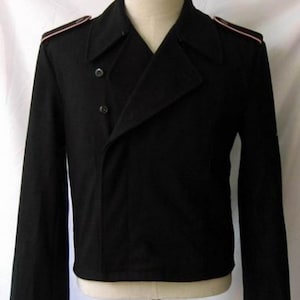 May include: A black double-breasted jacket featuring a wide collar, long sleeves, and shoulder epaulets with thin red piping. The garment has two visible dark buttons on the front closure and is displayed on a mannequin torso.