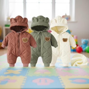 Op de afbeelding: Drie baby-sneeuwpakken in bruin, groen en crème. Elk pak heeft een capuchon met berenoren, een gewatteerd patroon en een berenpatch op de borst. De pakken hebben een ritssluiting aan de voorkant en zijn voor baby's.
