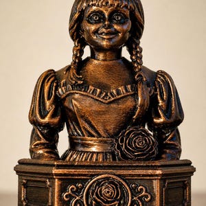 May include: A bronze-colored bust of a doll with braided hair, wearing a dress with a rose detail. The bust sits on a decorative base with a rose emblem. The doll has a slightly unsettling expression.