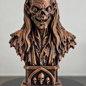 May include: A copper-colored bust of a gaunt, long-haired creature with a skeletal face and pointed ears. The figure wears a tattered, collared garment. The base features an arched alcove containing three small skulls in a row, finished in copper.