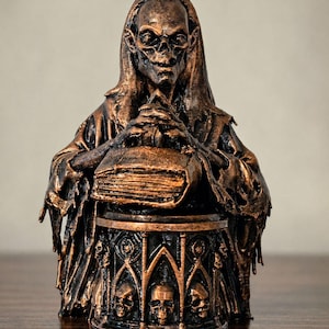 May include: A bronze-colored statue of a skeletal figure in a hooded robe, holding a book. The statue rests on a cylindrical base adorned with skulls and gothic arch designs. The figure's face is detailed with hollow eye sockets and a sinister grin.