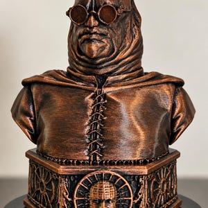 May include: A copper-colored bust of a figure wearing round goggles and a hooded garment with a central lace-up detail. The base is hexagonal, featuring a central circular relief of a face surrounded by a wheel-like design and chain motifs.