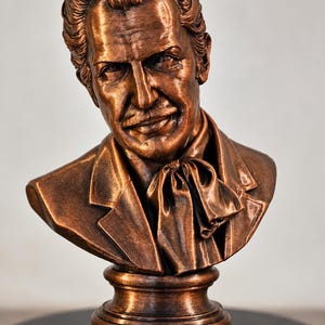 May include: A copper-toned bust sculpture depicting a man with wavy hair, a mustache, and a cravat. The figure wears a collared jacket and is mounted on a circular pedestal base. The finish has a metallic sheen with dark highlights.