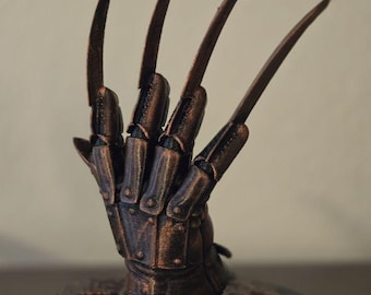 May include: A bronze-colored, metallic-looking glove sculpture featuring four long, sharp, blade-like fingers extending from a segmented hand piece. The object is mounted on a circular base with a textured, stone-like surface finish.