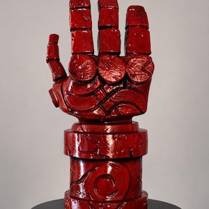 May include: A glossy red sculpture of a stylized, oversized hand with segmented fingers and a thick, cylindrical wrist cuff. The surface features intricate, carved circular and linear patterns throughout the entire structure, creating a textured appearance.