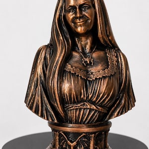 May include: A bronze-colored bust of a person with long hair, wearing a dress with a lace-trimmed neckline and a pendant necklace. The base of the sculpture features a repeating relief pattern of bats with wings spread, finished in a dark metallic tone.