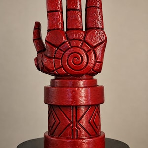 May include: A bright red, stylized sculpture of a large, blocky hand with a prominent spiral design on the palm. The wrist section features a thick, cylindrical base decorated with geometric diamond patterns and vertical lines in a matching red finish.