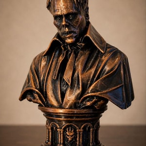 May include: A bronze-colored bust of a figure with a gaunt face, wearing a collared coat with a necktie. The figure rests on a circular pedestal base featuring architectural arch details and a small central skull emblem with a metallic finish.