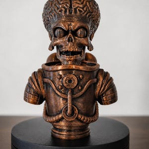 May include: A bronze-colored bust of an alien character with a large, exposed brain, skeletal facial features, and wide, dark eyes. The figure wears a detailed, textured suit with mechanical elements, including circular chest plates and ribbed shoulder sections.