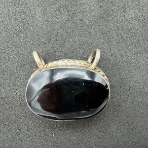 May include: An oval-shaped pendant featuring a smooth, polished black stone set in a silver-toned metal frame. The top of the pendant has two small, rounded loops for attachment, and the metal border displays a textured, patterned surface.
