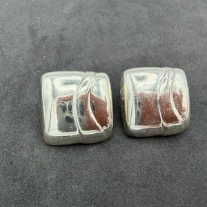 May include: A pair of square-shaped silver-toned earrings with a polished, reflective surface. Each earring features a smooth, rounded square silhouette with a subtle, raised diagonal ridge detail across the side, creating a clean, metallic look.