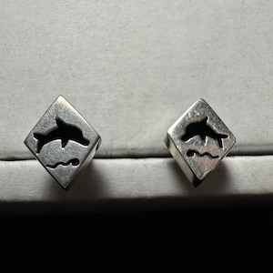 May include: A pair of silver-toned metal stud earrings, each shaped as a diamond-like cube featuring a cutout silhouette of a jumping dolphin above a small wavy line representing water, with a polished metallic finish.