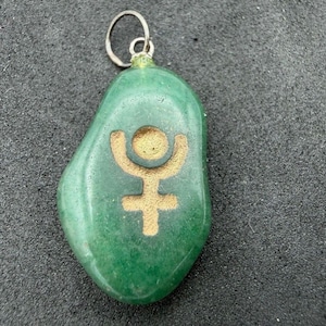 Puede incluir: Un colgante de piedra verde pulida y lisa con el símbolo planetario de Plutón grabado en color dorado en la parte frontal. El colgante cuenta con una sencilla argolla de metal plateado en la parte superior para colgarlo.