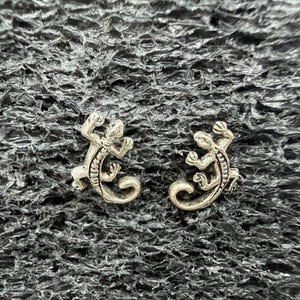 May include: A pair of silver-toned metal stud earrings shaped like small lizards. Each earring features a detailed, textured body with a curved tail and four legs, designed to resemble a gecko or similar reptile in a climbing pose.