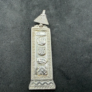May include: A silver-toned rectangular pendant featuring a pyramid-shaped bail at the top. The pendant displays a vertical column of five distinct embossed circular motifs, surrounded by a decorative border with geometric patterns.