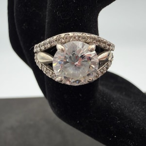 May include: A silver-toned ring featuring a large, round, clear faceted center stone held by four prongs. The band is split into two arched sections, each encrusted with a row of small, clear, round stones that frame the central setting.