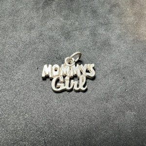 May include: A silver-toned metal charm featuring the text "MOMMY'S Girl" in a stylized, raised font. The charm includes a small circular jump ring at the top for attachment, with a polished, reflective surface finish throughout the piece.