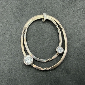 May include: A silver-toned pendant featuring two interlocking oval frames with a twisted metal texture. The pendant is accented with two round, clear, faceted stones set in circular bezels, positioned at the bottom and side of the frame.