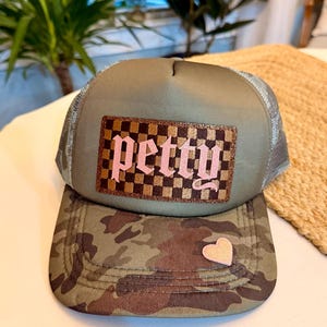 May include: A camouflage trucker hat with a pink heart and the word "petty" in pink script on a checkered patch. The hat has a mesh back and a curved brim.