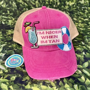 May include: A bright pink baseball cap with a beige mesh back. The front features an embroidered patch of a blue cocktail with a straw and orange slice, a rectangular patch with the text I'M NICER WHEN IM TAN, and a blue and white life preserver.