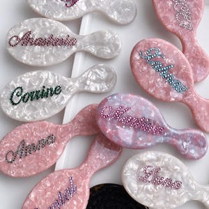 May include: A collection of personalized hairbrushes in various colors, including pink and white, with names spelled out in sparkling rhinestones. The brushes have an oval shape with a handle and are arranged on a white tray.