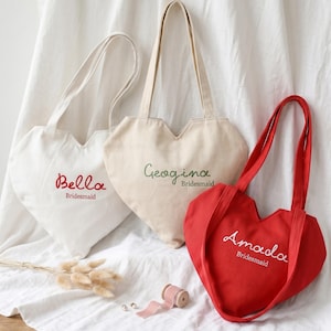 Può includere: Tre borse tote a forma di cuore, bianche, beige e rosse. Ogni borsa ha un lungo manico e testo ricamato: "Bella", "Georgina" e "Amada" con "Bridesmaid" sotto. Le borse sono su un tessuto bianco.