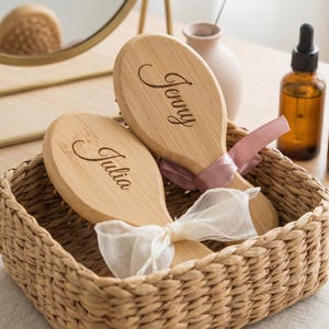 May include: Two wooden hairbrushes, each engraved with a name, "Jenny" and "Julia", sit in a woven basket. The brushes are adorned with ribbons. A mirror and a bottle are visible in the background.