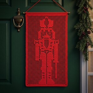 Op de afbeelding: Een rechthoekige rode gehaakte wandhanger met een gedetailleerd silhouet van een notenkraker, gemaakt met opengewerkte rastersteken. Het stuk hangt aan een horizontale roede met een dun rood koord en toont een uniform, ingewikkeld garenpatroon.
