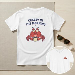 May include: A white t-shirt featuring a graphic of a red crab holding a steaming mug with the text CRABBY IN THE MORNING printed above it. The back of the shirt displays the large graphic, while the front chest area shows a smaller version of the crab.