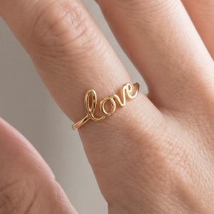 以下が含まれることがあります： 連続した筆記体でloveという単語が書かれたゴールドトーンのリング。金属製のバンドは細く滑らかで、l、o、v、eの文字が繋がった流れるようなデザインで指の上に配置されています。