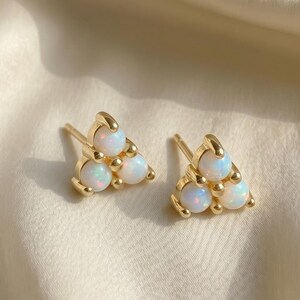 May include: A pair of gold stud earrings, each featuring three small, round, iridescent white opals arranged in a triangular cluster. The polished gold metal settings hold the stones securely, creating a delicate and symmetrical design for the ears.