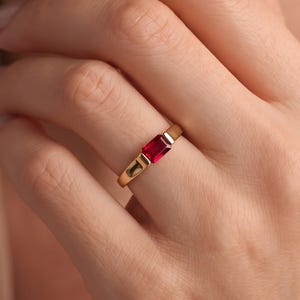 Puede incluir: Un anillo de oro con una piedra preciosa roja rectangular de corte esmeralda, engastada horizontalmente en el centro de una banda de oro lisa y pulida. La piedra roja vibrante está sujeta de forma segura por un engaste de bisel integrado.