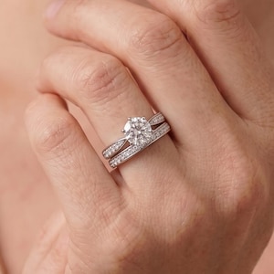Puede incluir: Un conjunto de anillos de compromiso en tono plateado con una piedra central redonda de talla brillante en un engaste de cuatro garras, acompañado de una alianza a juego. Ambos anillos tienen pequeñas piedras en pavé a lo largo de los aros.