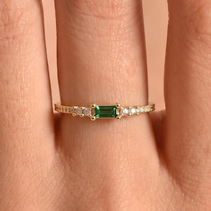 Könnte beinhalten: Ein goldener Ring mit einem zentralen, rechteckigen, smaragdgrünen Edelstein im Baguetteschliff, der auf jeder Seite von drei kleinen, runden, klaren Steinen flankiert wird. Das Band ist dünn und metallisch mit einem polierten Finish.