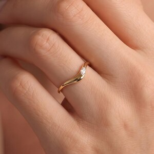 May include: A gold ring featuring a delicate, wavy band design with a single, small, clear marquise-cut gemstone set off-centre. The polished metal band curves gently, creating a fluid, asymmetrical silhouette that rests on the finger.