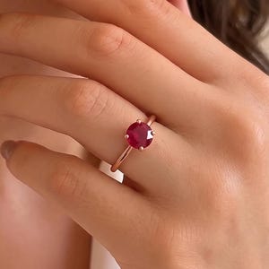 May include: A ring featuring a round, faceted, deep red gemstone set in a polished rose gold band. The stone is secured by four prongs, showcasing a vibrant crimson hue that contrasts with the warm metallic tone of the slender, circular band.