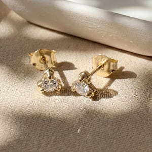 May include: A pair of gold stud earrings, each featuring a single round clear gemstone held in a three-prong setting. The earrings have a polished gold finish and are designed with a post back for secure attachment to the earlobe.