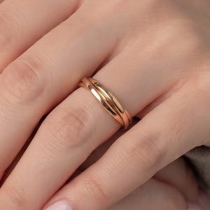 Op de afbeelding: Een roségouden ring met een ontwerp van drie banden met gladde, gepolijste oppervlakken. De ring wordt om de vinger gedragen en toont een doorlopende, onderling verbonden metalen structuur met een warme, metallic afwerking.