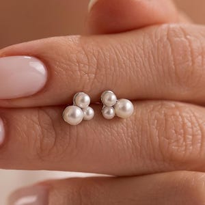 May include: A pair of stud earrings featuring a cluster of three small white pearls arranged in a triangular formation. The pearls are set on a metallic base, creating a compact and rounded design that sits flush against the earlobe when worn.