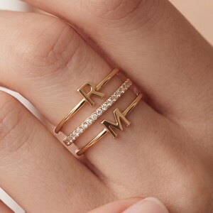 Puede incluir: Un conjunto de tres anillos de tono dorado usados juntos. El anillo superior presenta la letra R, el anillo central es una banda delgada incrustada con pequeñas piedras claras y el anillo inferior presenta la letra M. Los anillos tienen un acabado pulido.