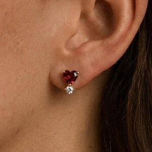 May include: A stud earring featuring a heart-shaped deep red gemstone positioned above a smaller, round, clear white gemstone. The earring is set in a metallic frame and is secured to the earlobe with a post and backing mechanism.