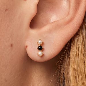 May include: A gold stud earring featuring a vertical arrangement of three small round stones. The design consists of a white pearl at the top, a black onyx stone in the center, and a white pearl at the bottom, all set in a thin gold metal bar.