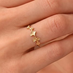 May include: A gold ring featuring a series of small, textured ginkgo leaf motifs arranged in a row along the band. The metal has a polished finish, and the design consists of multiple connected leaf shapes that wrap around the finger.